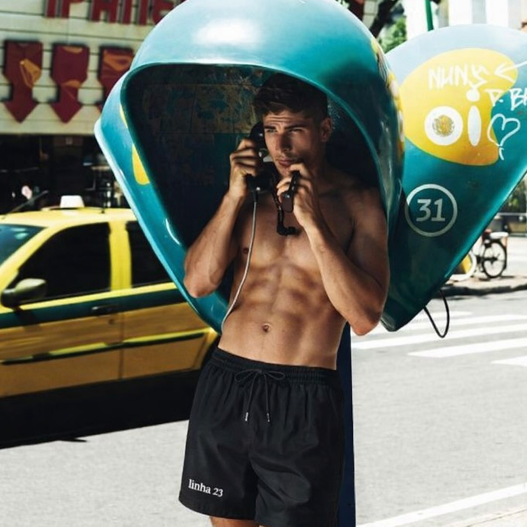 Shirtless male model wearing black Brazil core swim shorts in an urban Rio-inspired street scene, representing Brazilian men's fashion style.